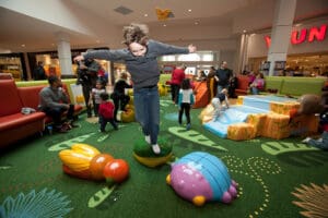 A lively indoor play area filled with children is seen, with a central focus on a young boy in a grey shirt and jeans jumping with arms spread wide on a green cushioned structure shaped like an insect. Adults and children are interacting in the background, showcasing how children learn through play.