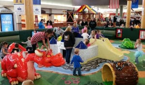 Children and adults are gathered at a vibrant indoor play area filled with colorful structures like a red dragon, a yellow slide, and a green crocodile. Within this soft play environment, children learn while parents supervise their fun. The surroundings feature playful, whimsical decor.
