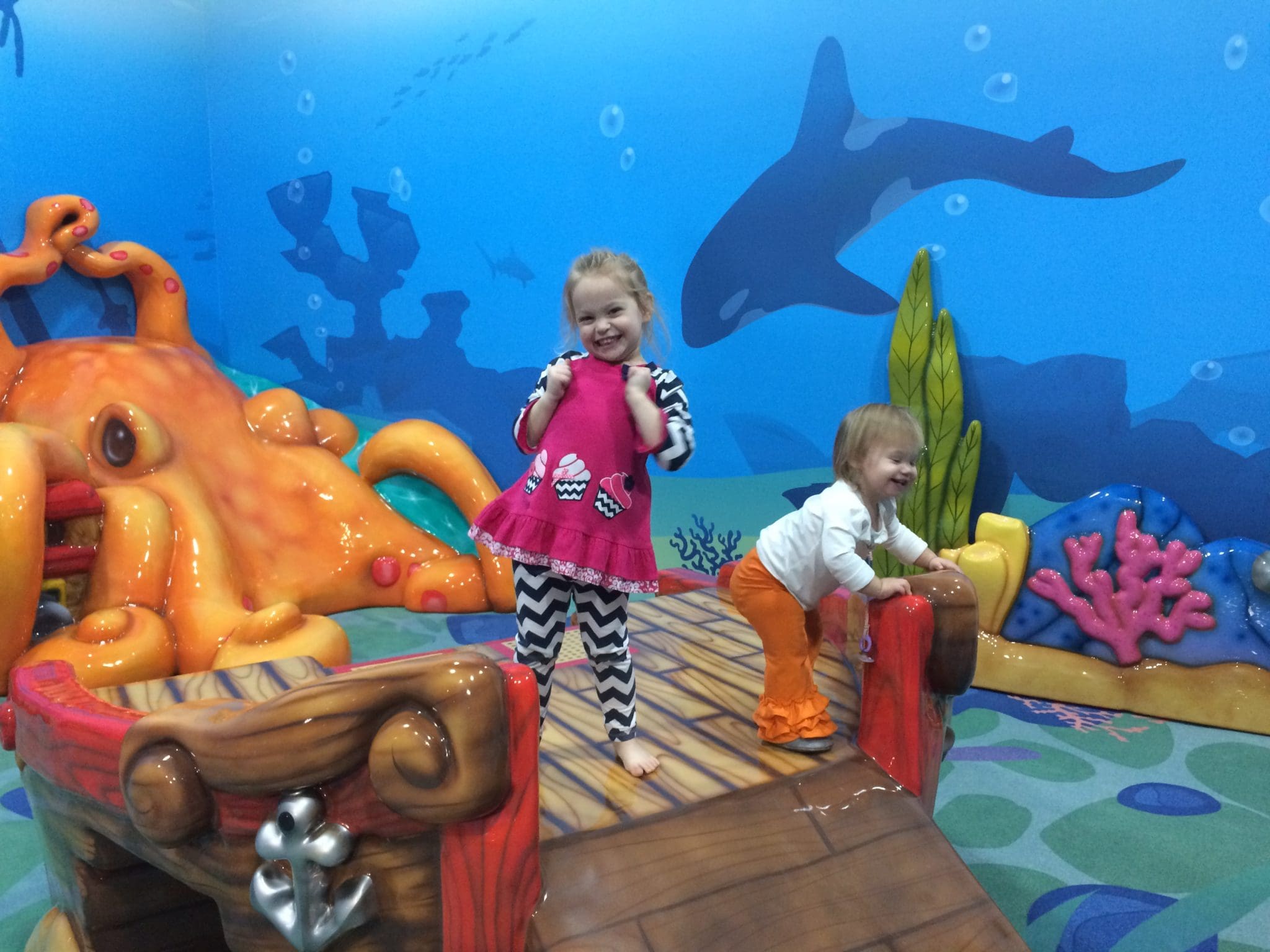 Two young children explore the aquatic-themed play space at a vibrant hotel resort. The girl, dressed in pink and patterned leggings, stands on a wooden bridge, while the younger boy in orange pants laughs nearby. Bright, colorful sea creature decorations and a blue ocean mural surround them.
