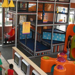 Children's indoor play area with multiple levels of climbing structures featuring colorful padded surfaces, a ball pit, and musical note decor hanging on the sides. Two kids are enjoying the experience on the equipment while others engage with interactive screens nearby.
